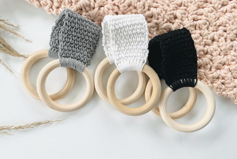 Puede incluir: Tres anillos de dentici&oacute;n de madera con correas de crochet de diferentes colores. Los anillos son de madera natural y las correas son grises, blancas y negras.
