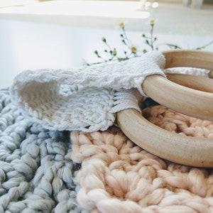 Puede incluir: Dos anillos de madera con una correa de crochet blanco, descansando sobre una manta de crochet gris y rosa.