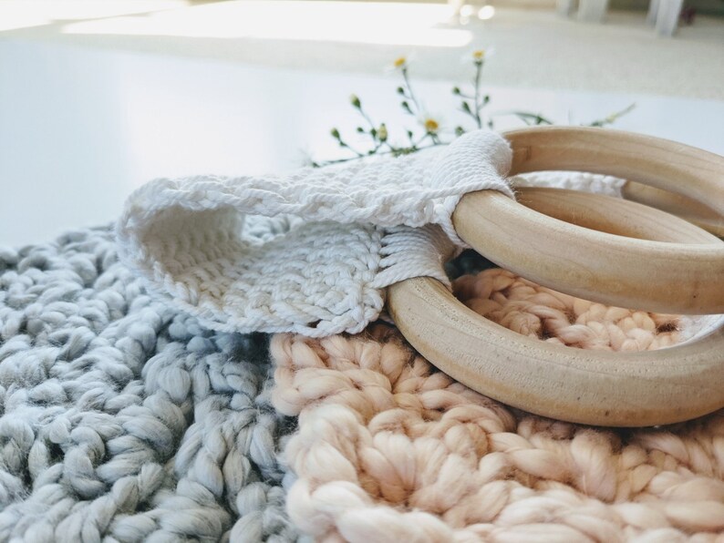 Puede incluir: Dos anillos de madera con una correa de crochet blanco, descansando sobre una manta de crochet gris y rosa.