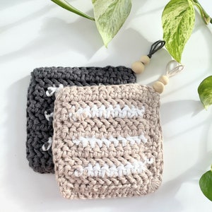 May include: Two crocheted dishcloths, one beige with white stripes and one gray, with wooden beads attached to the top.