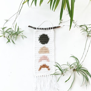 May include: White crocheted wall hanging with a green circle, tan, pink, and orange triangles. The hanging has a white fringe at the bottom.