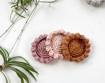 Puede incluir: Tres posavasos de ganchillo en diferentes colores: rosa, marrón claro y gris. Los posavasos son redondos y tienen un borde festoneado.