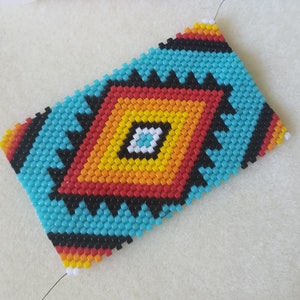 Puede incluir: Un primer plano de una pulsera de cuentas con un patrón geométrico en tonos de azul, rojo, amarillo, naranja y negro. La pulsera está hecha con cuentas pequeñas y redondas y está enhebrada en un hilo delgado.