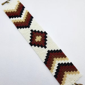 May include: A white beaded bracelet with a geometric pattern in brown, black, and beige. The bracelet has a metal clasp.