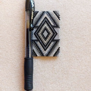 May include: A black and white beaded pen holder with a geometric pattern. The pen holder is next to a black Pilot G-2 pen.