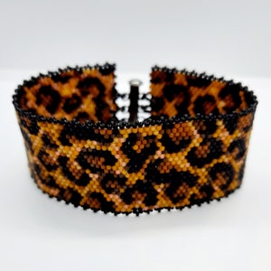 May include: A beaded bracelet with a leopard print pattern in shades of brown, black, and pink. The bracelet is made with small, round beads and has a metal clasp.