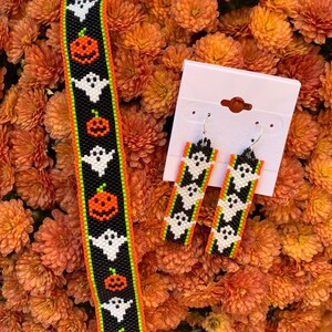 Op de afbeelding: Een set Halloween-sieraden van kralen met een zwart en groene armband met witte geesten en oranje pompoenen, en een paar bijpassende oorbellen met hetzelfde ontwerp.