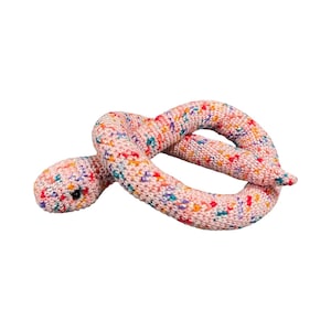 May include: A crocheted snake toy in a light pink color with colorful flecks. The snake is coiled and has a small black eye. The yarn has a speckled pattern with red, blue, yellow, and purple dots.