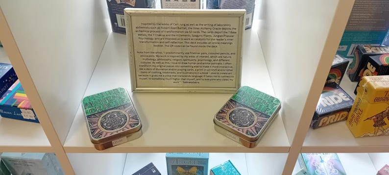 Puede incluir: Dos barajas de cartas de juego verdes y doradas con un dise&ntilde;o en blanco y negro en la parte delantera. Las barajas est&aacute;n en un estante blanco con otras barajas de cartas de juego. Una descripci&oacute;n enmarcada de las barajas est&aacute; encima de las barajas.