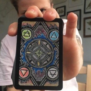 Puede incluir: Una carta negra con un dise&ntilde;o colorido que presenta cuatro c&iacute;rculos con s&iacute;mbolos en su interior. Los s&iacute;mbolos son un tri&aacute;ngulo con una l&iacute;nea que lo atraviesa, un c&iacute;rculo con una l&iacute;nea que lo atraviesa, un tri&aacute;ngulo con una l&iacute;nea que lo atraviesa y un c&iacute;rculo con una l&iacute;nea que lo atraviesa. Los c&iacute;rculos est&aacute;n dispuestos en un c&iacute;rculo alrededor de un dise&ntilde;o central.