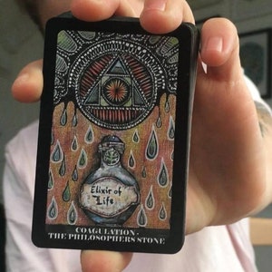 Puede incluir: Una carta negra con una ilustraci&oacute;n colorida de un tri&aacute;ngulo con un ojo en su interior, rodeado de l&iacute;quido que gotea. La carta tambi&eacute;n presenta una botella etiquetada como "Elixir of Life" y el texto "Coagulation - The Philosopher's Stone".