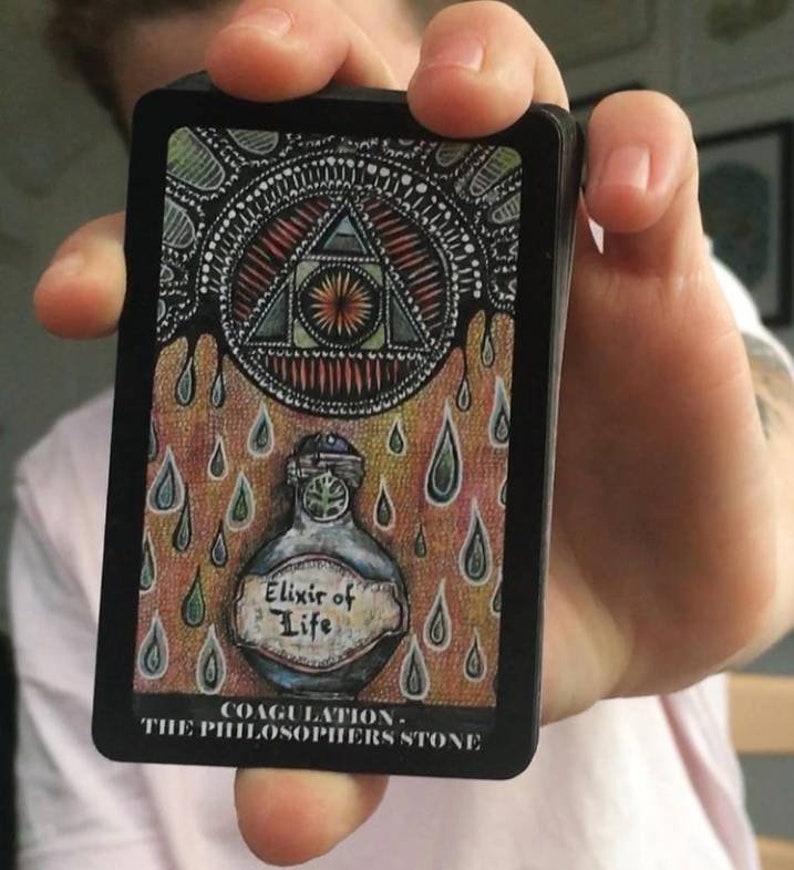 Puede incluir: Una carta negra con una ilustraci&oacute;n colorida de un tri&aacute;ngulo con un ojo en su interior, rodeado de l&iacute;quido que gotea. La carta tambi&eacute;n presenta una botella etiquetada como "Elixir of Life" y el texto "Coagulation - The Philosopher's Stone".