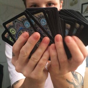Puede incluir: Una persona est&aacute; sosteniendo una baraja de cartas con un dise&ntilde;o azul, verde y amarillo. Las cartas tienen un s&iacute;mbolo de tri&aacute;ngulo en ellas.