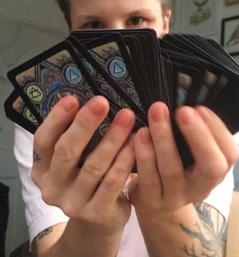 Puede incluir: Una persona est&aacute; sosteniendo una baraja de cartas con un dise&ntilde;o azul, verde y amarillo. Las cartas tienen un s&iacute;mbolo de tri&aacute;ngulo en ellas.