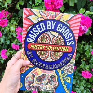 Puede incluir: Una colección de poesía titulada "RAISED BY GHOSTS" de Sabrakadabra. La portada del libro presenta una colorida ilustración de calavera, diseños florales y un emblema circular azul con el título. El fondo incluye flores rosas vibrantes.