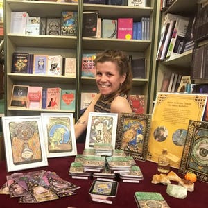 Puede incluir: Una mujer se sienta frente a una estanter&iacute;a, mostrando una variedad de art&iacute;culos a la venta, incluyendo un juego de cartas de or&aacute;culo, peque&ntilde;as cajas de hojalata y cristales. Las cartas de or&aacute;culo est&aacute;n enmarcadas y presentan ilustraciones coloridas. Las cajas de hojalata est&aacute;n apiladas en forma de pir&aacute;mide. Los cristales est&aacute;n dispuestos en una peque&ntilde;a pila. El texto en el cartel dice "Inner Alchemy Oracle by Babes Kadabra".