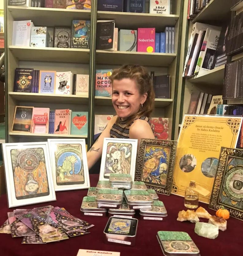 Puede incluir: Una mujer se sienta frente a una estanter&iacute;a, mostrando una variedad de art&iacute;culos a la venta, incluyendo un juego de cartas de or&aacute;culo, peque&ntilde;as cajas de hojalata y cristales. Las cartas de or&aacute;culo est&aacute;n enmarcadas y presentan ilustraciones coloridas. Las cajas de hojalata est&aacute;n apiladas en forma de pir&aacute;mide. Los cristales est&aacute;n dispuestos en una peque&ntilde;a pila. El texto en el cartel dice "Inner Alchemy Oracle by Babes Kadabra".