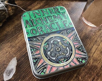 Könnte beinhalten: Eine grüne und goldene Metallbox mit den Worten "Inner Alchemy Oracle" auf der Vorderseite. Die Box hat ein farbenfrohes Mandala-Design mit einem Farbschema in Pink, Gelb und Blau.