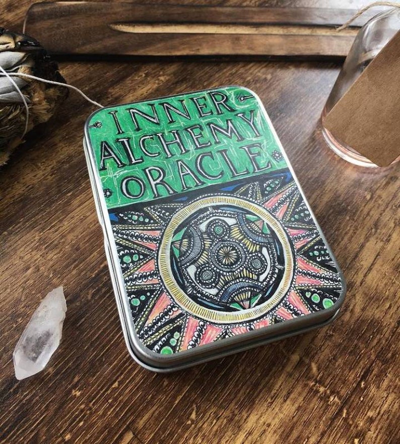Puede incluir: Una caja de metal verde y dorada con las palabras "Inner Alchemy Oracle" impresas en la parte delantera. La caja tiene un dise&ntilde;o de mandala colorido con un esquema de color rosa, amarillo y azul.