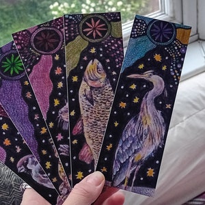 Puede incluir: Conjunto de seis marcapáginas coloridos con ilustraciones de diferentes animales. Cada marcapáginas tiene un fondo negro con un diseño colorido y un patrón de estrellas. Los animales incluyen un pez, una garza, un gato, un pájaro, una flor y una mariposa.