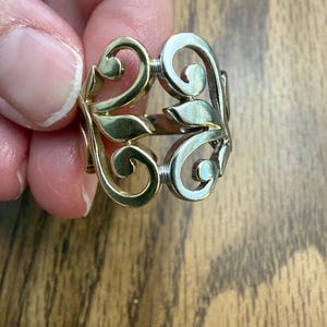 May include: A two-tone ring featuring a polished gold-colored metal section and a silver-colored metal section. The design consists of openwork scroll and leaf-like motifs that form a continuous band around the finger, showcasing a smooth finish.