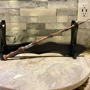 Może przedstawiać: Czarna i srebrna różdżka z fakturowanym uchwytem jest prezentowana na czarnym stojaku. Różdżka spoczywa na białym marmurowym podłożu.