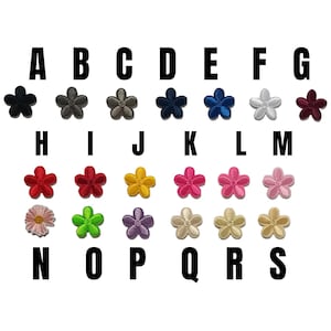 Puede incluir: Una colección de diecinueve pequeños parches bordados con forma de flor dispuestos en tres filas, etiquetados de la A a la S. Los parches presentan varios colores sólidos, incluyendo negro, gris, marrón, azul, blanco, rojo, amarillo, rosa, verde, morado y dorado.