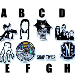 Könnte beinhalten: Eine Sammlung von gestickten Aufnähern mit Figuren und Symbolen aus einer gothik-thematischen Fernsehserie. Die Aufnäher zeigen eine Figur, die Geige spielt, eine Silhouette eines Mädchens, eine Figur mit einem Regenschirm, ein Familienporträt, eine Hand, ein Spinnennetz und den Text "SNAP TWICE".