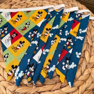 May include: A fabric bunting banner featuring triangular flags with a repeating Mickey Mouse print. The design includes a diamond pattern in yellow, red, and green, alternating with solid blue sections decorated with white Mickey Mouse silhouettes.