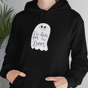 May include: Black hooded sweatshirt with a white ghost graphic and the text "I'm here for the Boos". The ghost has a smiling face. The sweatshirt has a front pocket and a drawstring hood. A fun Halloween-themed apparel item.