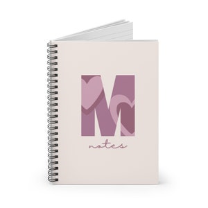 May include: A spiral-bound notebook with a light pink cover. The cover features a large letter "M" in shades of purple, with heart shapes incorporated into the design. The word "notes" is written below the letter in a cursive font.
