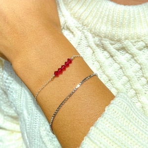 Może przedstawiać: Srebrna bransoletka z łańcuszkiem i rzędem czerwonych kryształowych koralików. Bransoletka jest na nadgarstku osoby.