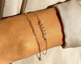 May include: Two gold bracelets on a person's wrist. One bracelet is a delicate chain with a row of clear crystal beads. The other bracelet is a thicker chain with a more substantial link.