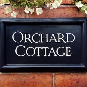 May include: A black rectangular sign with the words "ORCHARD COTTAGE" in white lettering. The sign is framed and mounted on a red brick wall, with greenery and white flowers at the top.