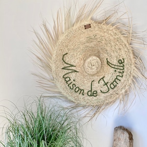 Op de afbeelding: Een ronde, geweven wanddecoratie met de tekst "Maison de Famille" in groene letters. Het heeft gedroogde palmbladeren en een klein leren label. Een groene plant en een stuk drijfhout staan op de voorgrond.