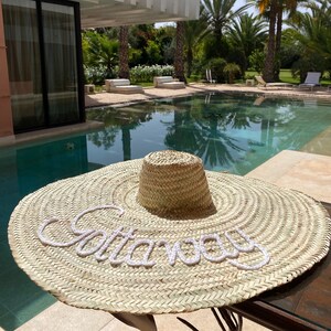 May include: A large, round, straw sun hat with a white embroidered word "Gatlinburg" on the brim. The hat is sitting on a table next to a swimming pool.