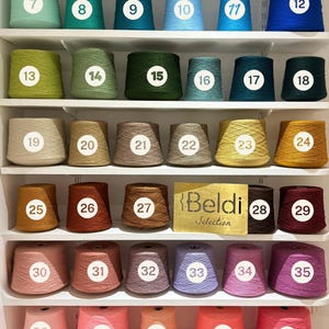 May include: A display of forty-six yarn cones arranged on white shelves, each labeled with a numbered white circle. The collection features a wide spectrum of colors including neutrals, blues, greens, yellows, browns, pinks, and reds. A gold sign reads Beldi Selection.