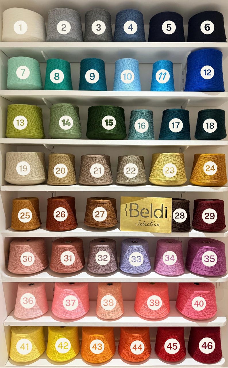 May include: A display of forty-six yarn cones arranged on white shelves, each labeled with a numbered white circle. The collection features a wide spectrum of colors including neutrals, blues, greens, yellows, browns, pinks, and reds. A gold sign reads Beldi Selection.