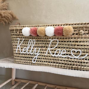 May include: A rectangular woven straw basket featuring a row of six decorative pom-poms in white, dusty rose, and tan colors. The front of the basket displays the name Kelly Rae in white cursive yarn lettering, ending with a small heart symbol.