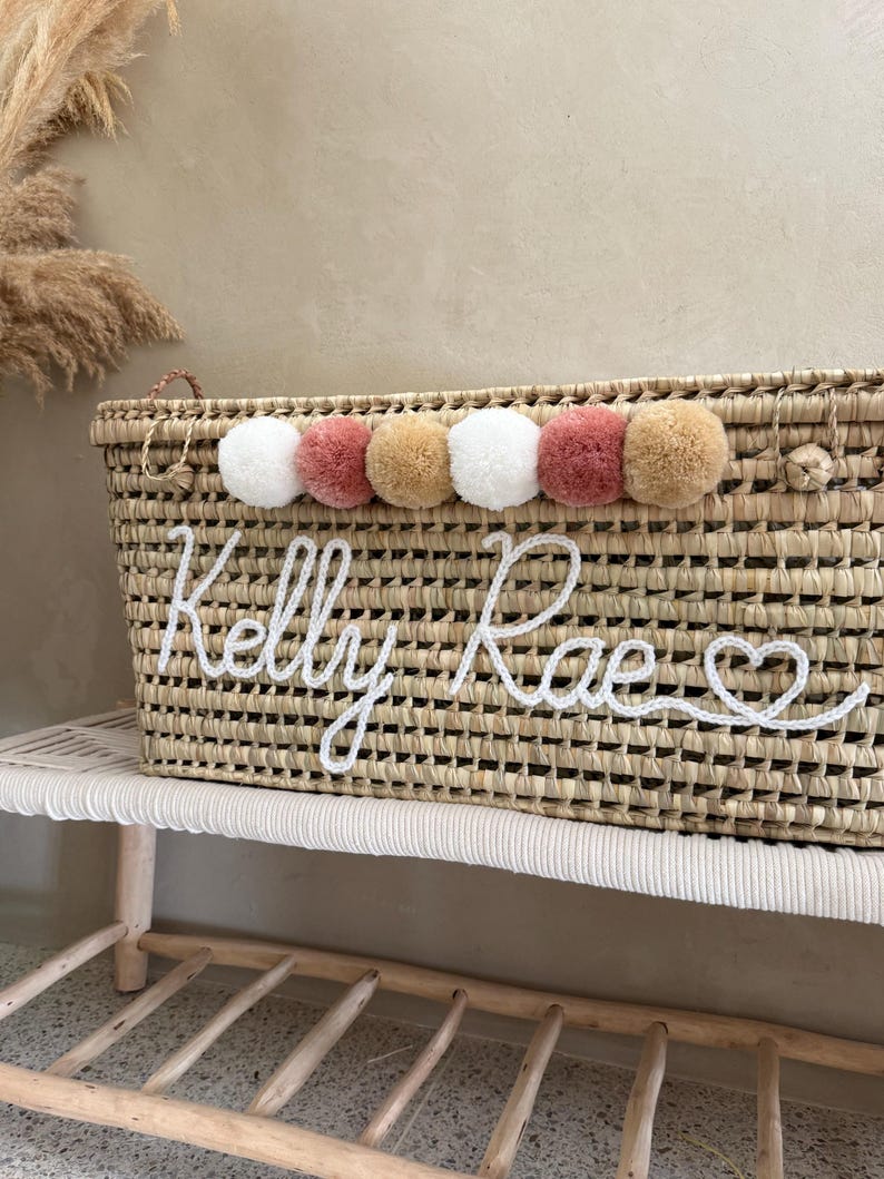 May include: A rectangular woven straw basket featuring a row of six decorative pom-poms in white, dusty rose, and tan colors. The front of the basket displays the name Kelly Rae in white cursive yarn lettering, ending with a small heart symbol.
