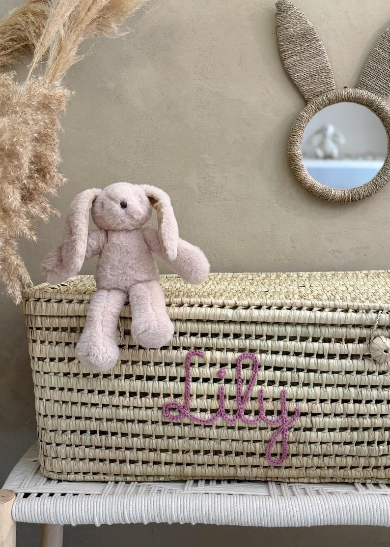 May include: A rectangular woven straw storage basket featuring the name Lily embroidered in purple yarn on the front. A soft, light pink plush rabbit toy with long floppy ears sits on the lid of the basket. The basket has a woven texture.