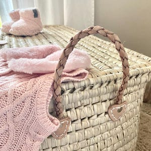 May include: A woven straw storage basket featuring a braided leather handle attached with heart-shaped leather patches. A soft pink cable-knit garment and a pair of fuzzy light pink booties are resting on top of the basket lid.
