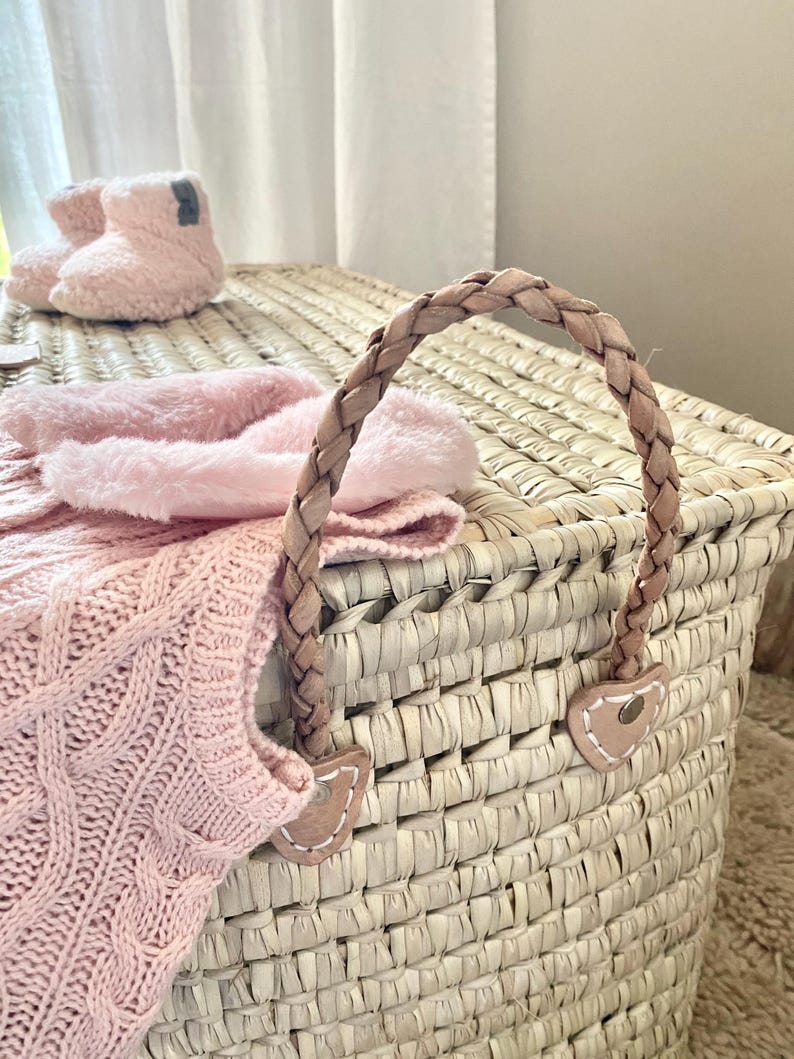 May include: A woven straw storage basket featuring a braided leather handle attached with heart-shaped leather patches. A soft pink cable-knit garment and a pair of fuzzy light pink booties are resting on top of the basket lid.