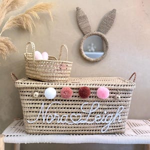 May include: A large rectangular woven straw basket with a lid, featuring the white embroidered name Nora Leigh, a heart symbol, and four colorful pom-poms. A smaller matching basket and a round mirror with woven straw bunny ears are included.
