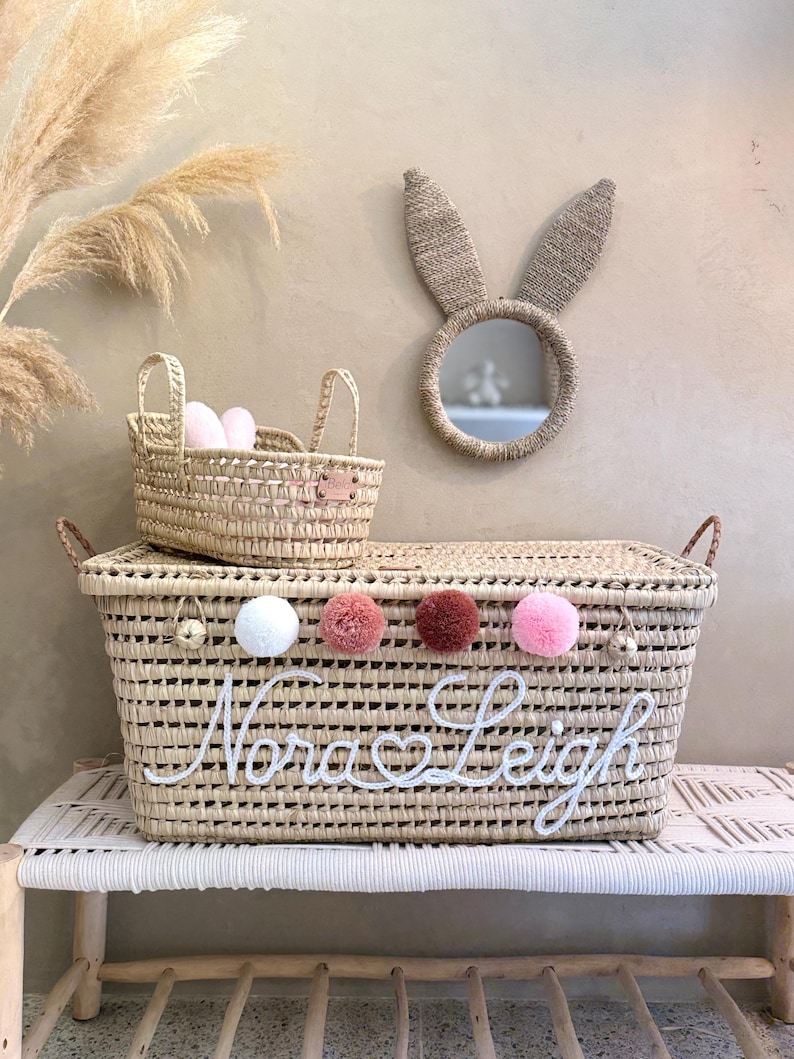 May include: A large rectangular woven straw basket with a lid, featuring the white embroidered name Nora Leigh, a heart symbol, and four colorful pom-poms. A smaller matching basket and a round mirror with woven straw bunny ears are included.