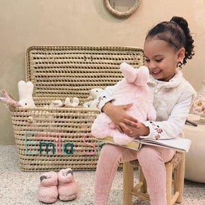 May include: A woven storage basket with a lid featuring the name Emilia Sofia embroidered in teal and pink. The basket contains several plush toys, including a white rabbit. A round mirror with woven rabbit ears hangs on the wall above.