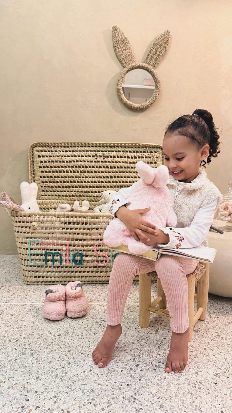 May include: A woven storage basket with a lid featuring the name Emilia Sofia embroidered in teal and pink. The basket contains several plush toys, including a white rabbit. A round mirror with woven rabbit ears hangs on the wall above.