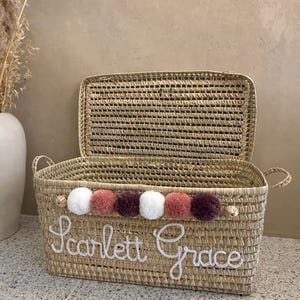 May include: A rectangular woven straw basket with a hinged lid and two side handles. The front features the white embroidered name Scarlett Grace and a row of six decorative pom-poms in alternating colors of white, dusty rose, and dark plum.