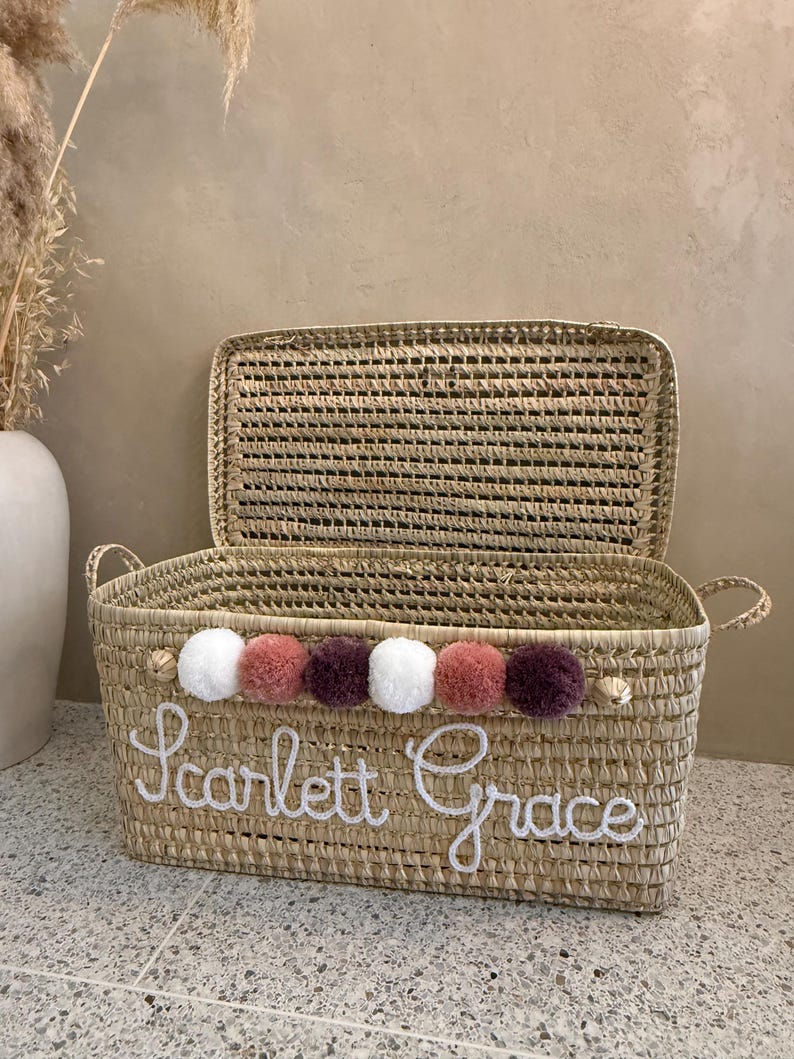 May include: A rectangular woven straw basket with a hinged lid and two side handles. The front features the white embroidered name Scarlett Grace and a row of six decorative pom-poms in alternating colors of white, dusty rose, and dark plum.