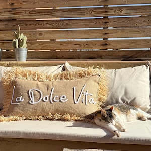 Pode incluir: Uma almofada de sofá bege com uma borda de franjas e o texto "La Dolce Vita" bordado em branco. Um gato está dormindo em um sofá branco com a almofada.